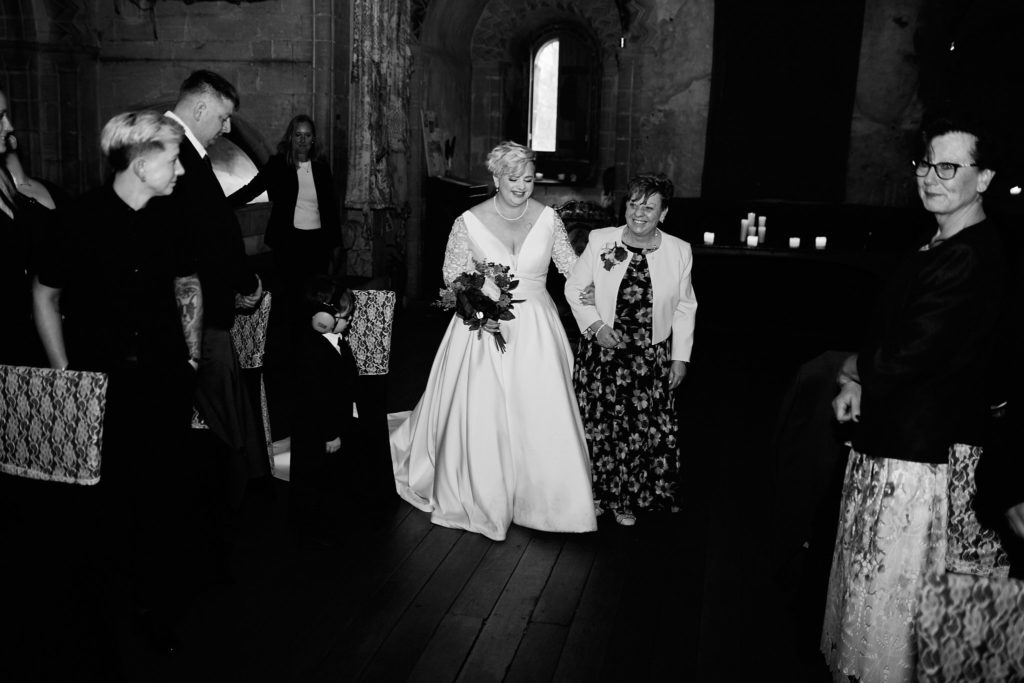 Black and white photo of the bride walking down the aisle arm in arm with a woman