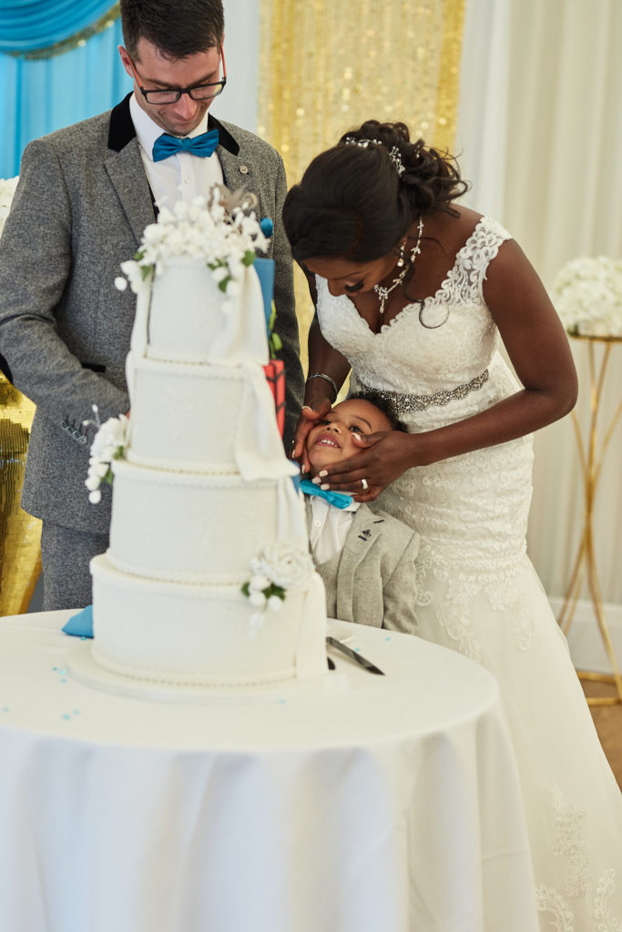 Bride laughing as she hugs a young page boy beside the wedding cake
