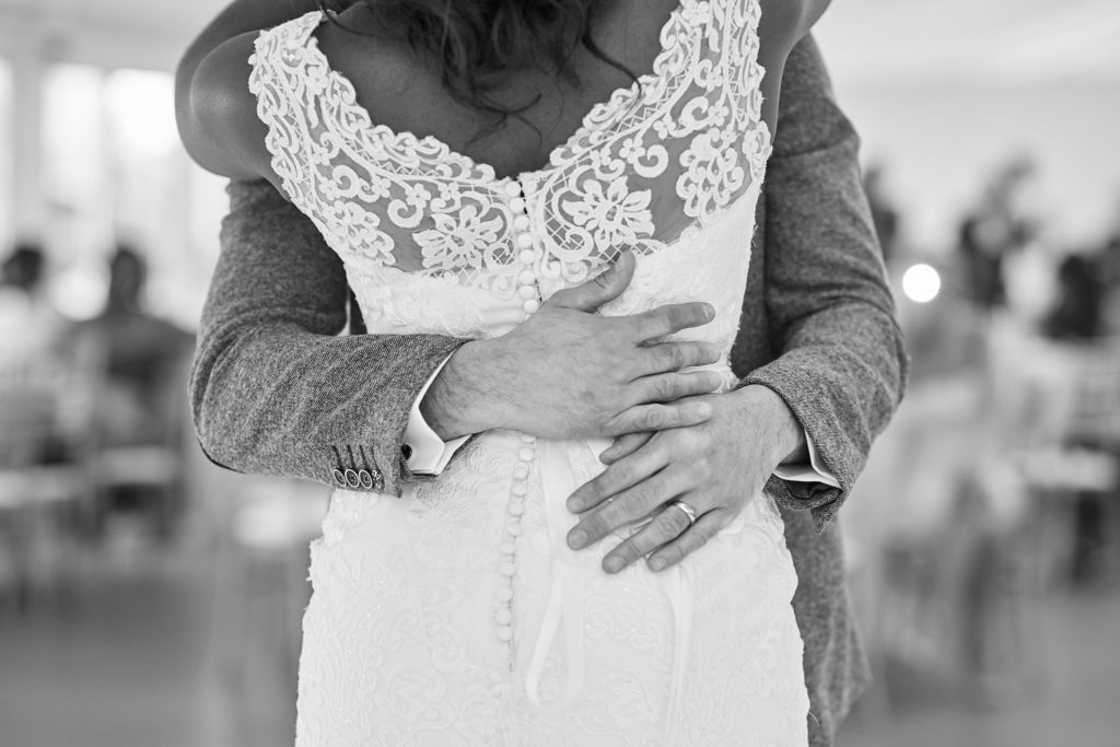 Black and white close-up of the groom's hands on the back of the bride's lace dress
