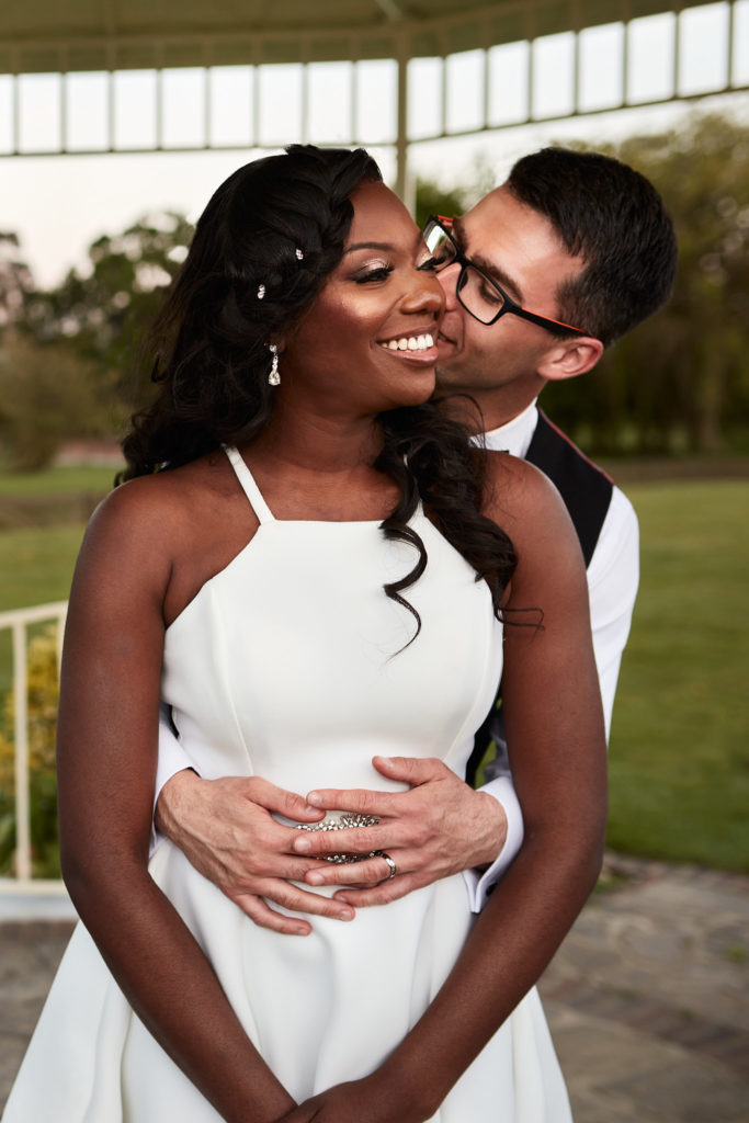 Groom hugging the bride from behind as she smiles in a white halterneck dress at Quendon Hall