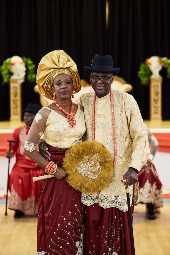 Older couple in traditional Itsekiri dress, the woman in a gold gele holding a woven fan