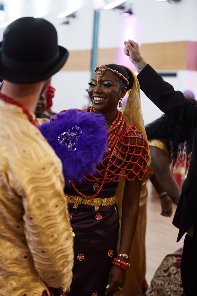 Bride laughing in a coral bead headpiece and cape, holding a purple feather fan