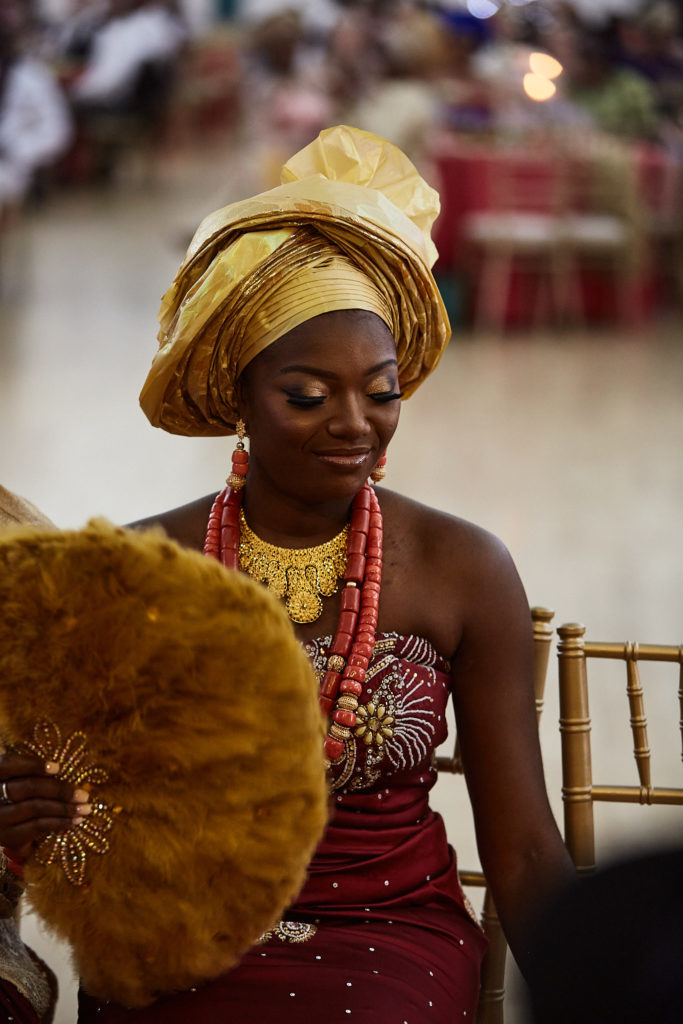 Bride smiling in a gold gele and coral beads with a woven fan