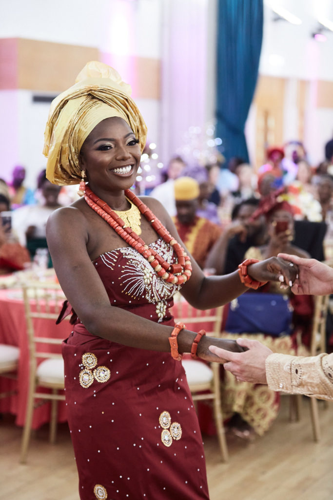 Bride smiling in a gold gele and red wrapper as she greets a guest