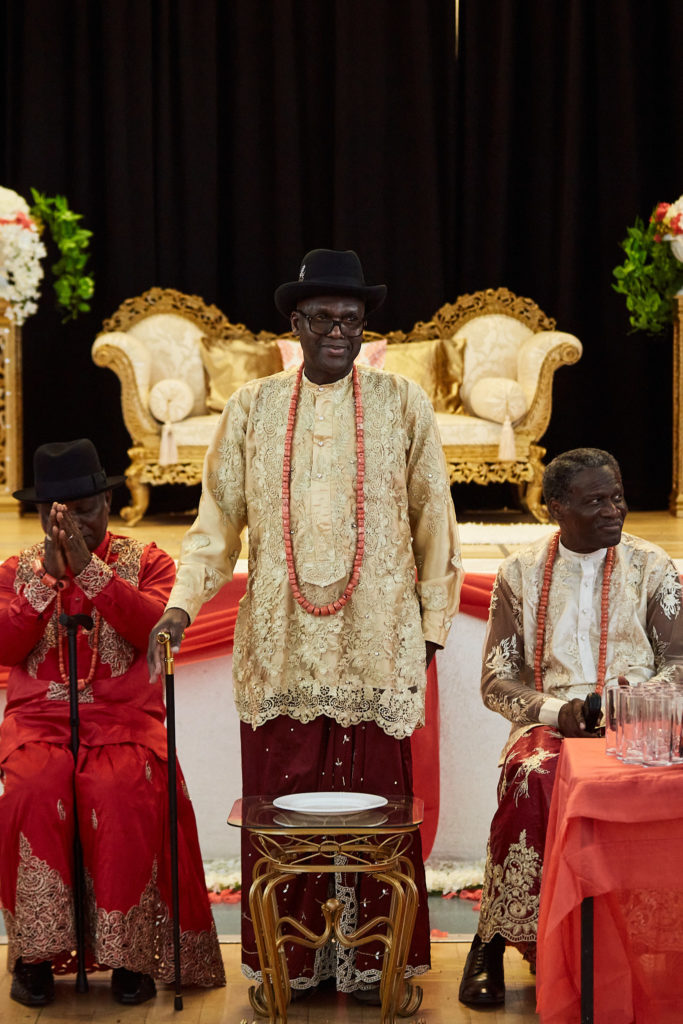 Three elders in traditional Itsekiri dress and hats seated in front of the gold sofa