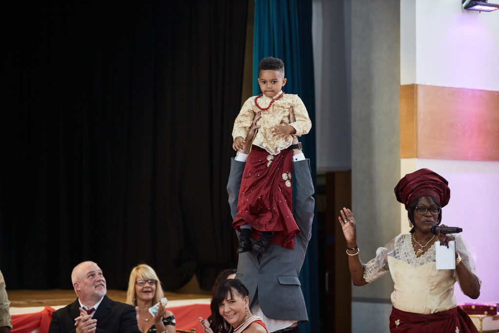 Young boy in traditional dress standing on a chair as a woman speaks with a microphone