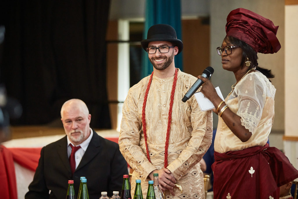 Groom standing beside a woman in traditional dress as she speaks with a microphone