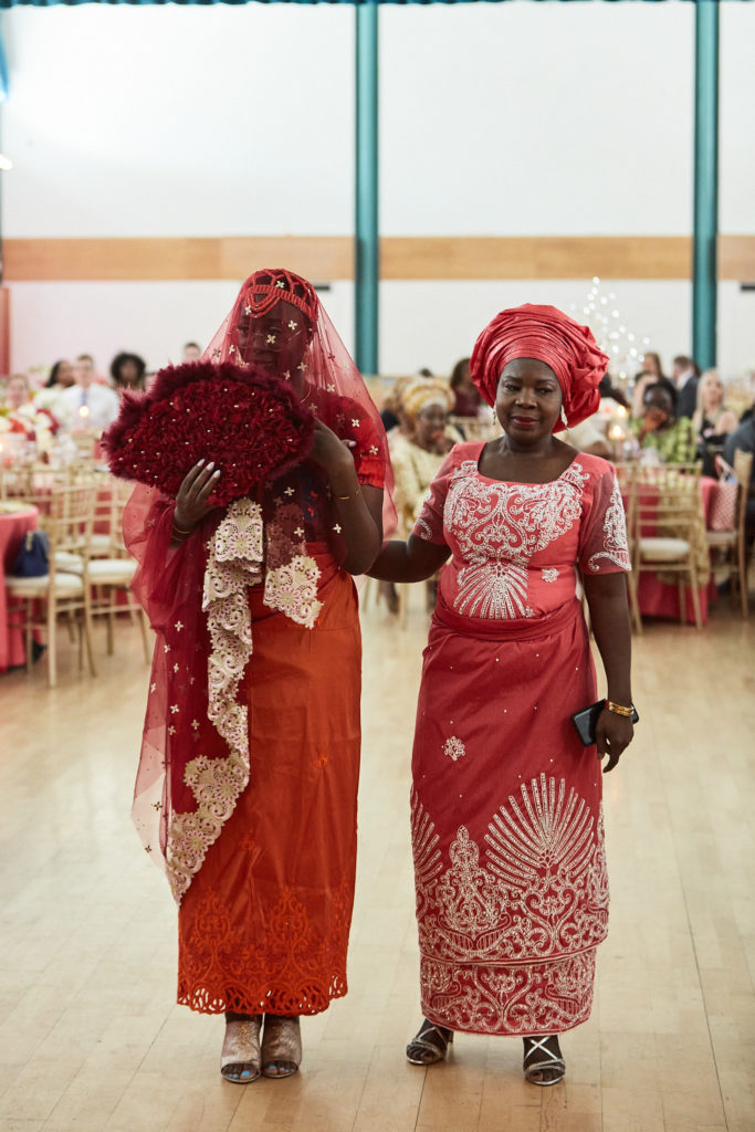 Bride in a red lace veil walking into the hall beside a woman in red