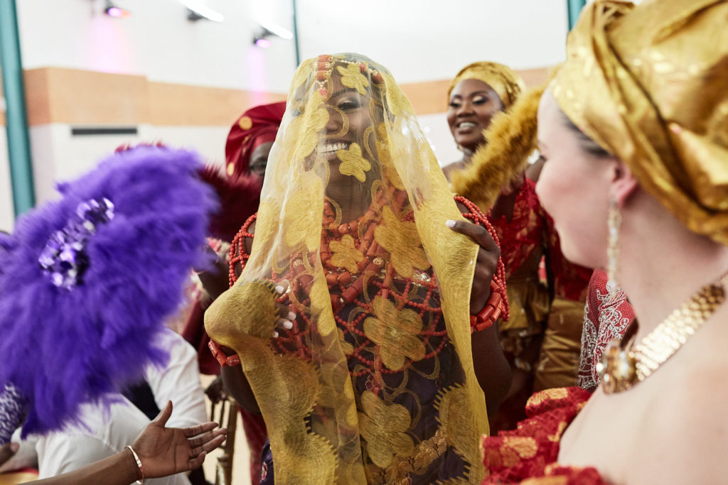 Bride under a gold lace veil among women holding a purple feather fan