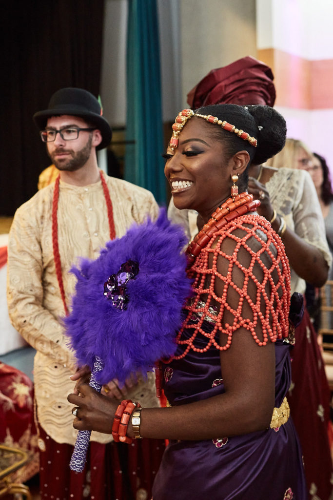 Bride laughing in coral beads beside the groom, holding a purple feather fan
