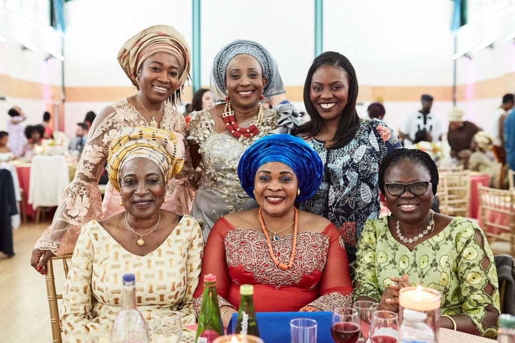 Six women in colourful traditional outfits and head ties smiling together at their table