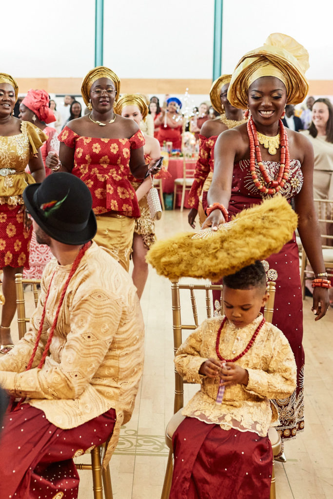 Bride in a gold gele and coral beads fanning a young boy in traditional dress as the groom sits beside him