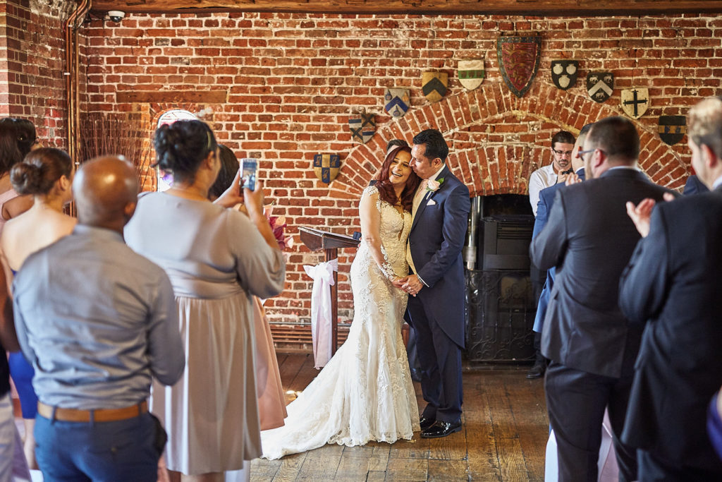 Bride and groom standing before their guests in the brick-walled ceremony room at Eltham Tudor Barn