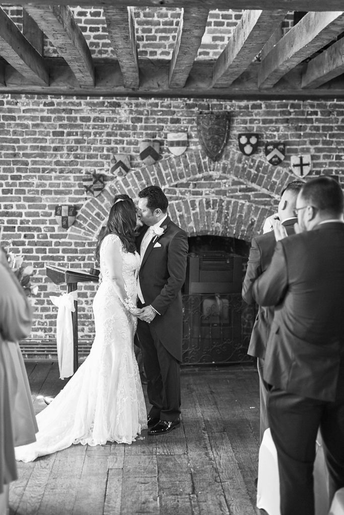 Black and white photo of the bride and groom kissing beneath the oak beams as guests watch