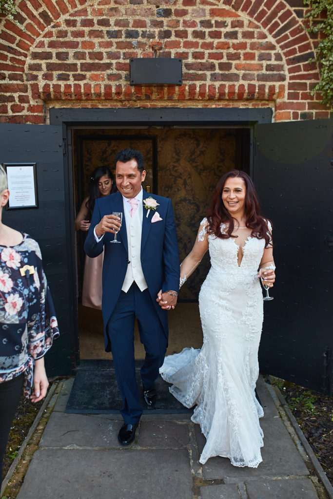 Bride and groom stepping out of Eltham Tudor Barn hand in hand, the groom raising a glass