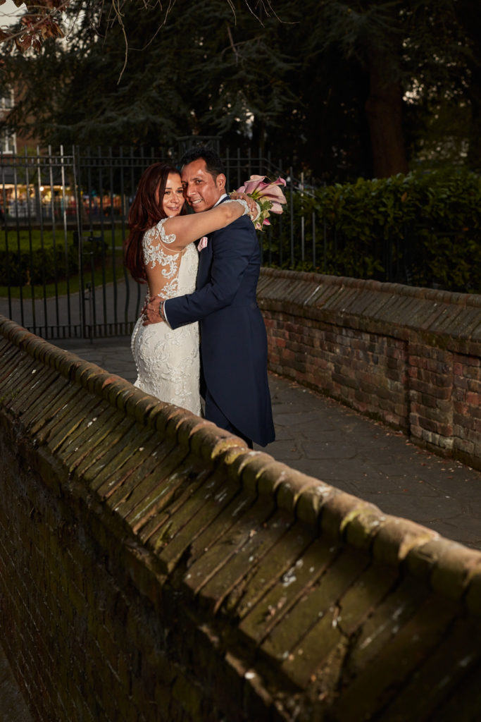 Bride and groom hugging beside a brick wall in the gardens at Eltham Tudor Barn