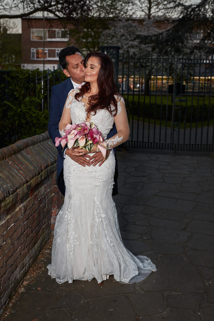 Groom kissing the bride's head as she holds her pink bouquet at Eltham Tudor Barn