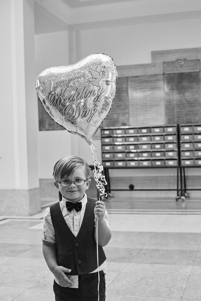 Black and white photo of a young page boy in a bow tie holding a heart-shaped balloon