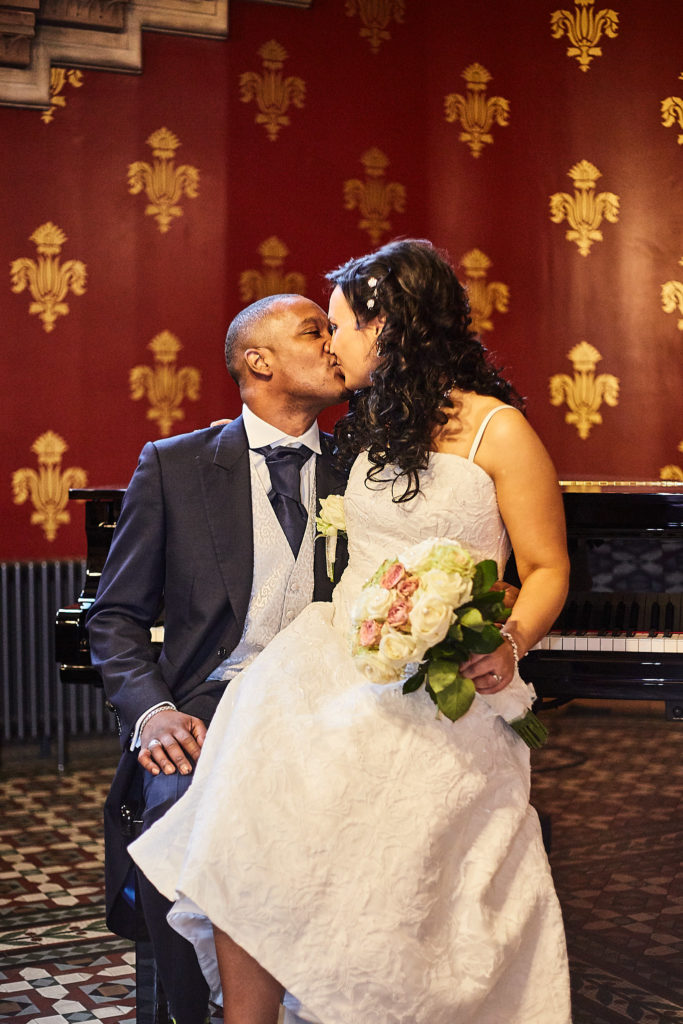 Bride kissing the groom beside a piano against red and gold wallpaper at the St Pancras Hotel