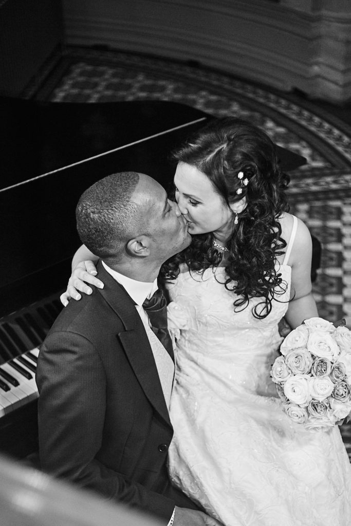 Black and white photo of the bride kissing the groom's cheek beside a piano at the St Pancras Hotel
