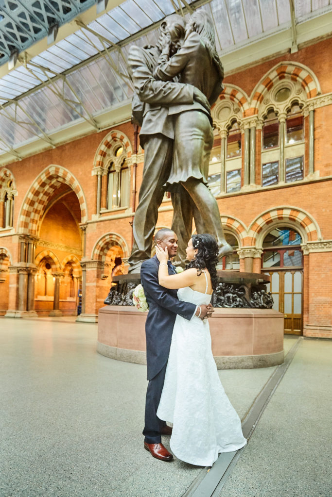 Bride and groom embracing beneath the Meeting Place statue at St Pancras station
