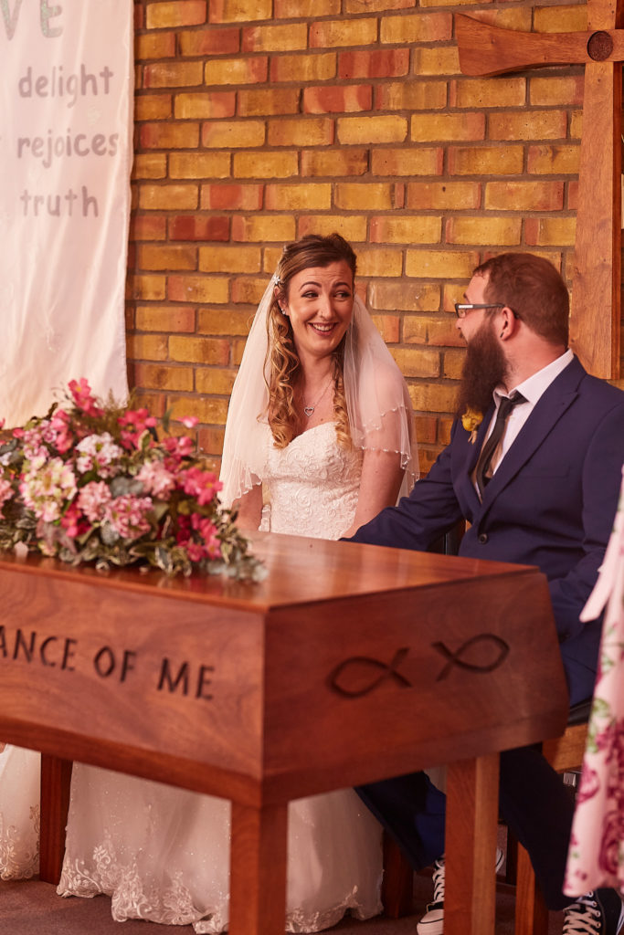 Bride and groom smiling as they sign the register beside a display of flowers