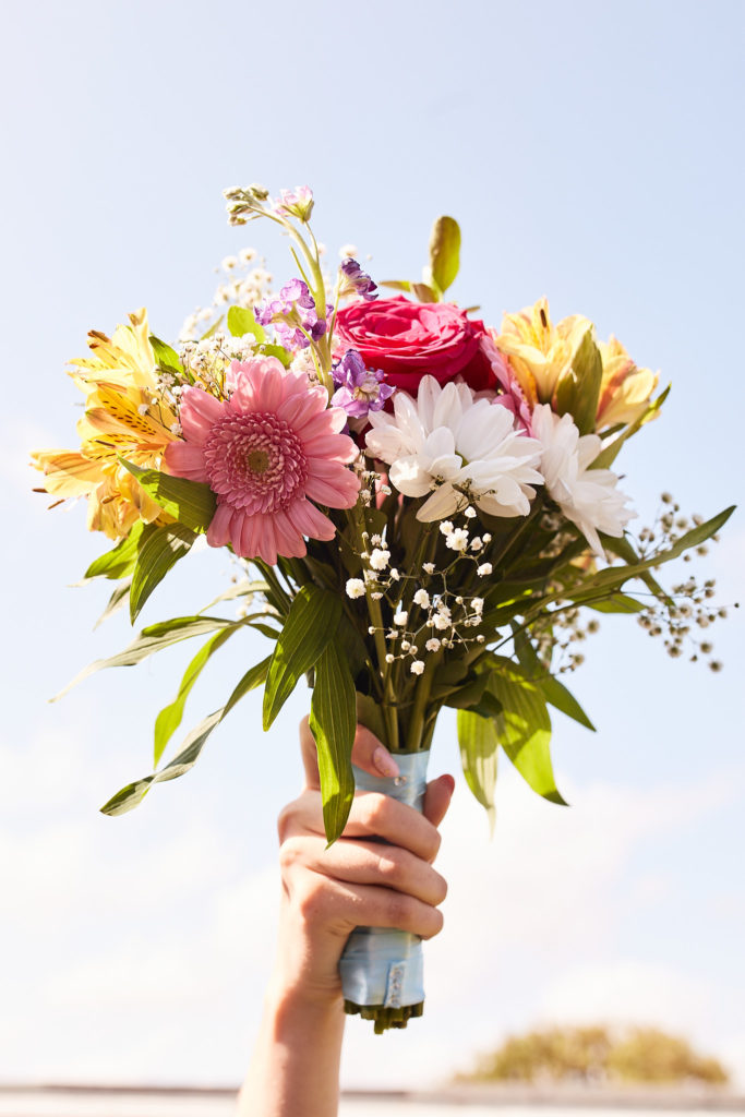 Hand holding a bouquet of pink, yellow and white flowers up against the sky