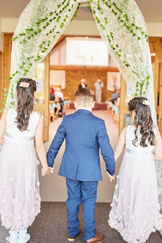 Page boy and two flower girls holding hands in the church doorway, seen from behind