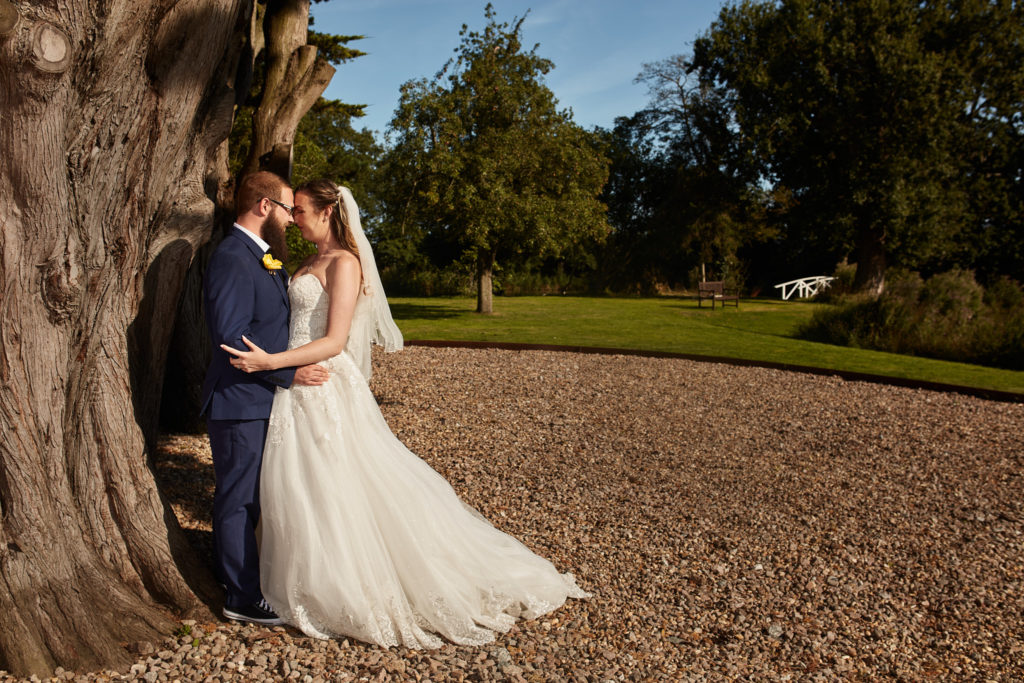 Bride and groom embracing beside a large tree in the grounds of Villiers Barn
