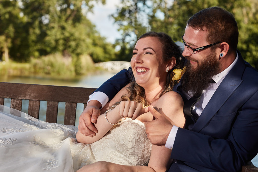 Bride laughing as she leans back against the groom by the lake at Villiers Barn