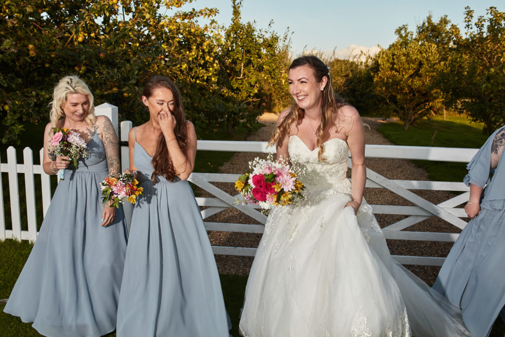 Bride laughing with bridesmaids in pale blue dresses beside a white gate