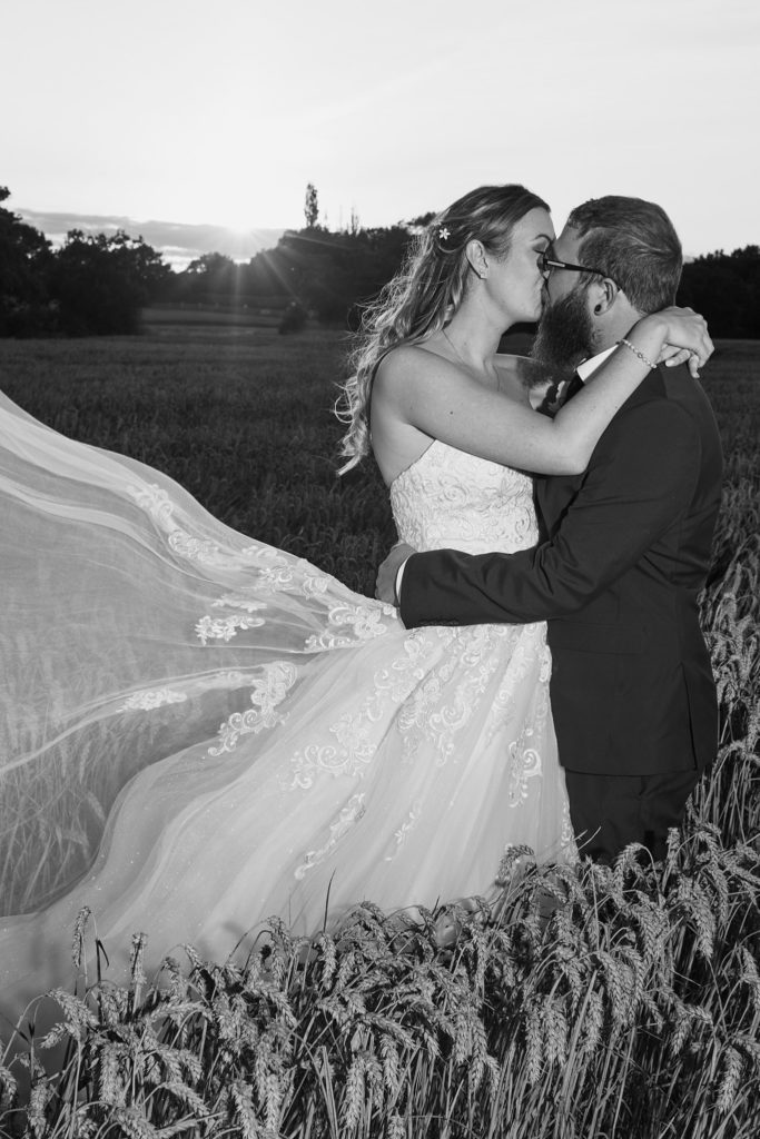 Black and white photo of the bride and groom kissing in a wheat field as her veil blows in the wind