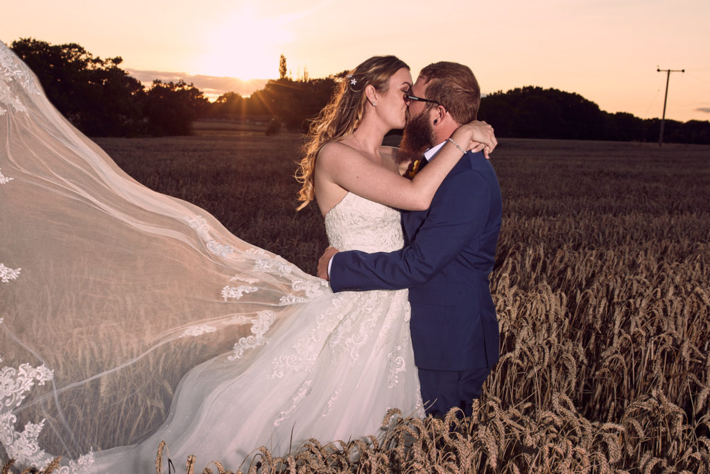 Bride and groom kissing in a wheat field at sunset with her veil flying, Villiers Barn