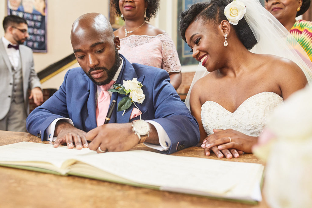 Groom signing the register as the bride smiles beside him