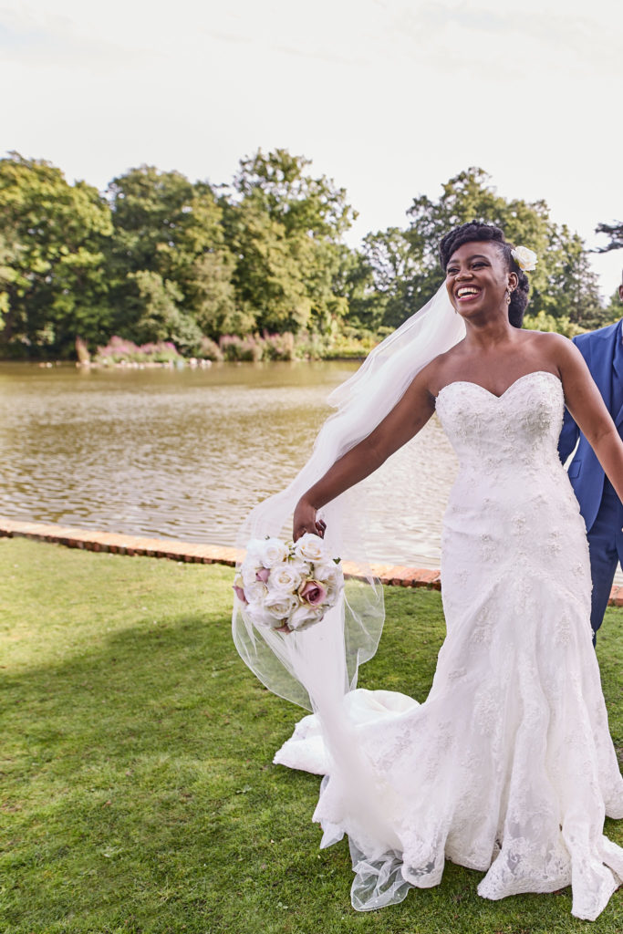 Bride laughing as she holds up her dress beside the lake at Forty Hall