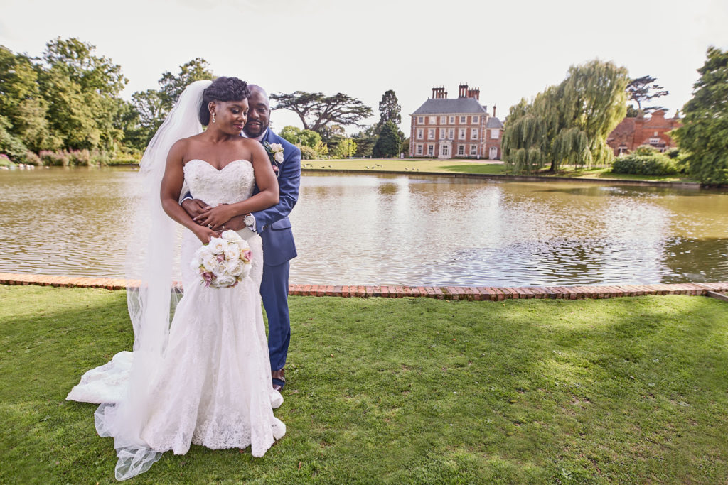 Groom hugging the bride from behind on the lawn with Forty Hall across the lake