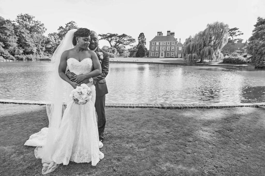 Black and white photo of the groom embracing the bride from behind by the lake at Forty Hall