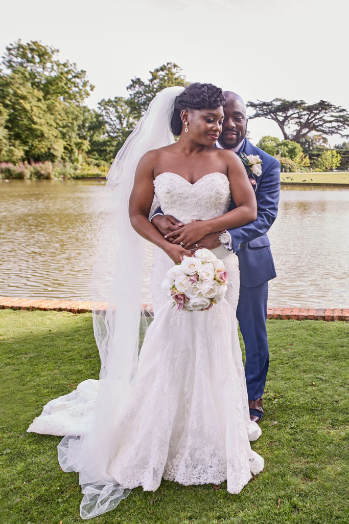 Groom holding the bride from behind as she smiles beside the lake at Forty Hall