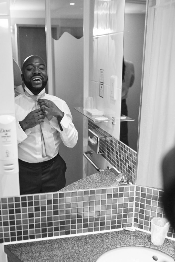 Black and white photo of the groom fastening his tie in a bathroom mirror