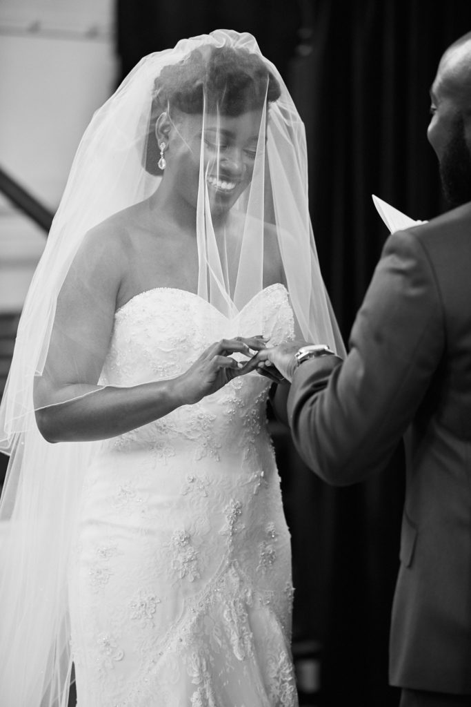 Black and white photo of the bride smiling beneath her veil as she holds the groom's hand