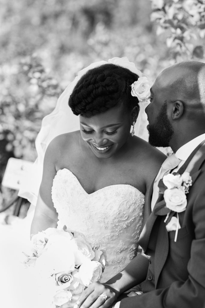 Black and white photo of the bride smiling down at her bouquet beside the groom
