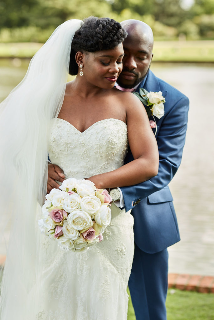 Groom holding the bride from behind as she looks down at her bouquet by the lake at Forty Hall