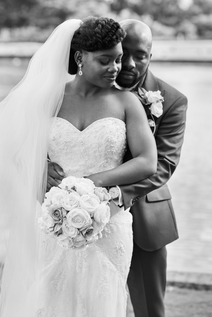 Black and white photo of the groom embracing the bride from behind beside the lake at Forty Hall
