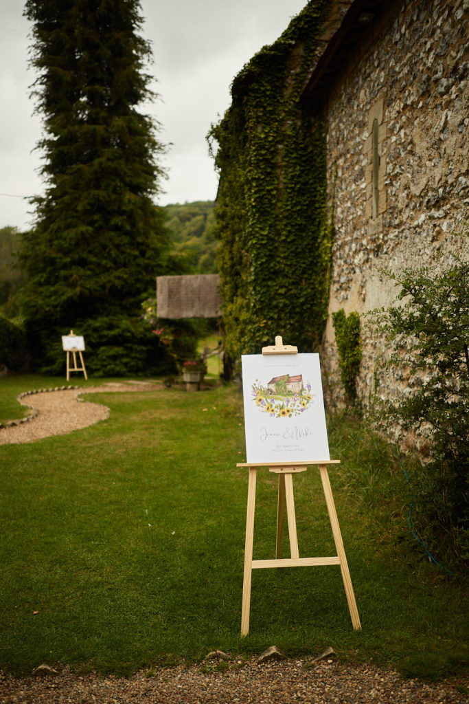 Wedding welcome sign on an easel beside an ivy-covered flint wall at the Lost Village of Dode