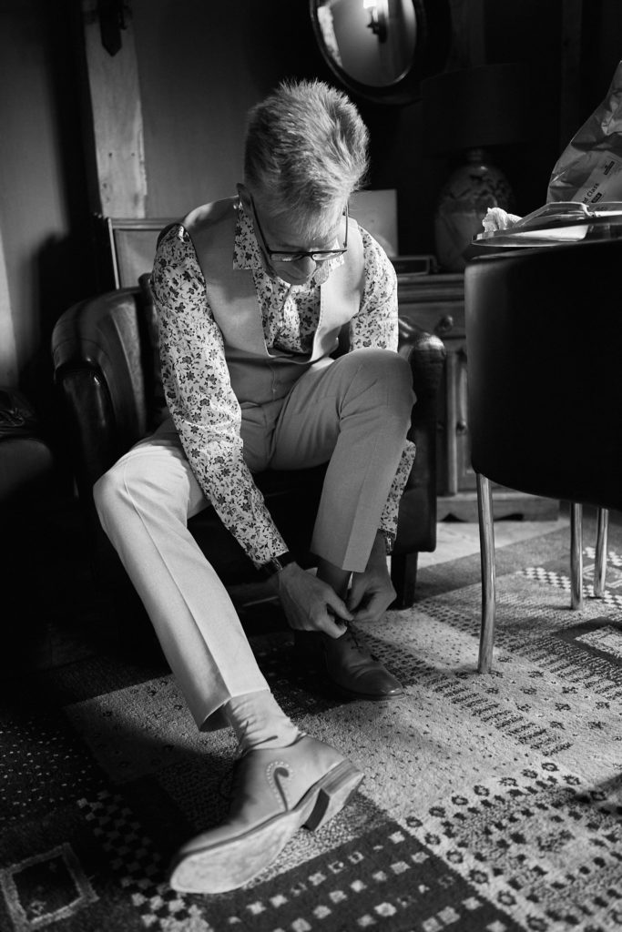 Black and white photo of the groom tying his shoe in a floral shirt and waistcoat