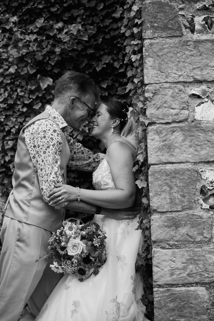 Black and white photo of the bride and groom forehead to forehead beside a flint wall at the Lost Village of Dode