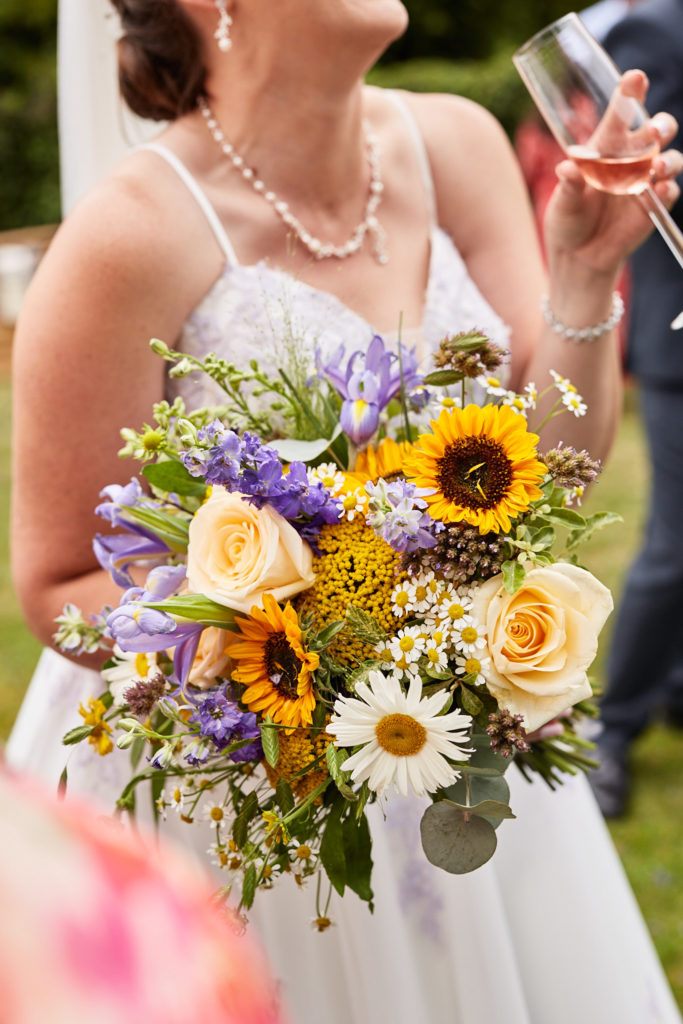 Bride holding a bouquet of sunflowers, daisies and roses with a glass of rosé