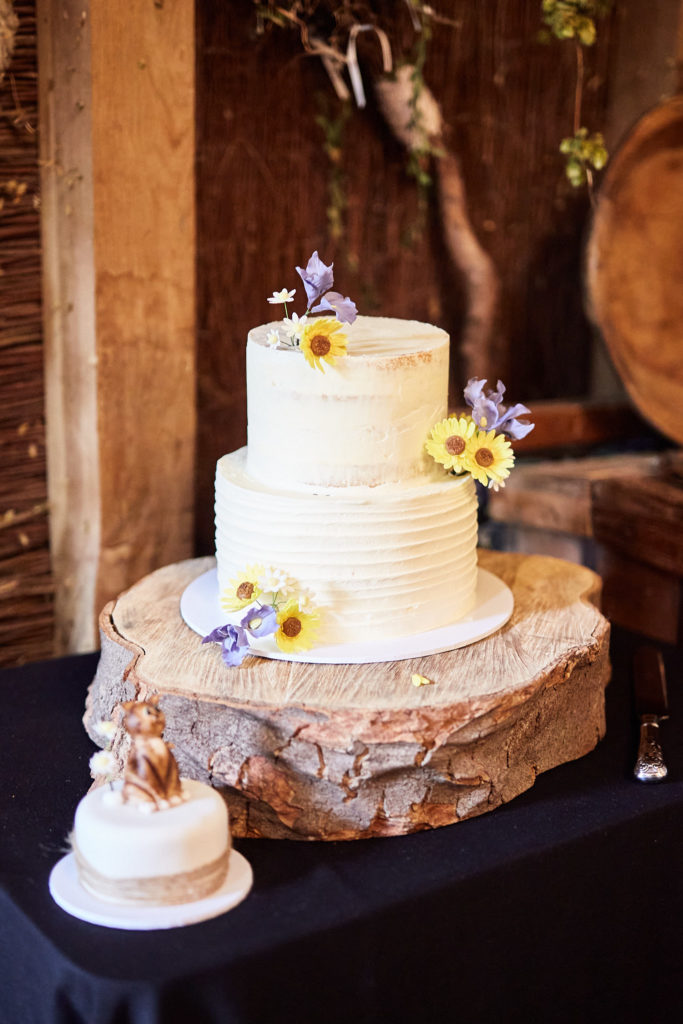 Two-tier wedding cake with sunflowers on a log slice, beside a small cake with a cat topper