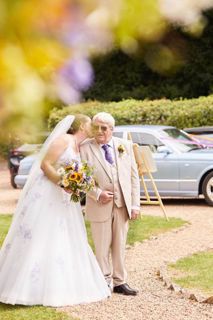 Bride walking arm in arm with an elderly man in a cream suit, a vintage car behind them