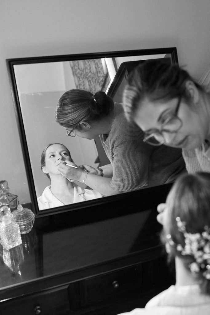 Black and white photo of the bride having her make-up done, reflected in a mirror